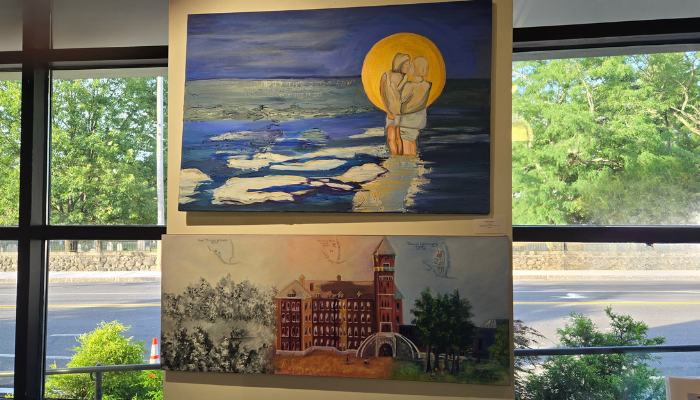 Two paintings on a wall, one of a couple in front of the moon and ocean at night and the other painting of a landscape photo with a building.